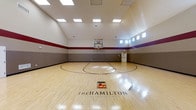 Located at The Hamilton Luxury Apartment Homes in Fishers, Indiana, this indoor basketball court is a standout amenity offering residents a professional-grade recreational facility. The court features a polished hardwood floor finished with regulation court markings, a prominent center-court logo bearing The Hamilton's branding, and a full basketball hoop and backboard setup. The space is defined by its impressively tall ceilings, which allow for unrestricted play and contribute to a spacious, open atmosphere. Recessed panel lighting is evenly distributed across the ceiling, ensuring consistent, well-lit conditions throughout the court at any time of day. High, horizontally-oriented windows line the upper walls, bringing in natural light while maintaining privacy and structural integrity. The walls are finished in a layered color scheme of gray tones accented by bold red and purple horizontal stripes, giving the court an energetic and polished aesthetic that reflects the community's upscale character. This single-floor facility is thoughtfully designed to meet the recreational needs of residents, combining functional athletic infrastructure with high-end finishes that align with the luxury standard of The Hamilton community in Fishers, IN.