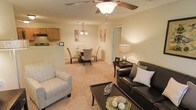 This single-floor residence showcases an open-concept design that seamlessly integrates the living, dining, and kitchen areas into one cohesive space. The living area is anchored by a ceiling fan with a light fixture overhead, providing both comfort and illumination. Warm beige carpeting extends throughout the main living spaces, contributing to the overall cozy atmosphere.

The living room accommodates a dark leather sofa paired with a patterned accent chair, centered around a dark wood coffee table. A console table with a table lamp adds warmth along one wall. The dining area sits just beyond, featuring a round table surrounded by upholstered chairs beneath a pendant chandelier, creating an intimate setting for meals.

The kitchen, partially visible beyond a raised breakfast bar, features light wood cabinetry and essential appliances. The raised bar serves as a natural divider between the kitchen and dining space while maintaining the open feel of the layout.

Warm, ambient lighting throughout the space is achieved through a combination of ceiling fixtures and accent lamps. Neutral wall tones and understated artwork contribute to a calm, welcoming atmosphere suited for everyday living.