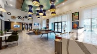 This single-floor clubhouse lounge is a thoughtfully designed social hub that blends eclectic style with practical amenity spaces. The main lounge area commands attention with soaring double-height ceilings accented by a teal-painted upper tier and a dramatic cluster of layered navy and gold pendant lights. Large-format light stone tile flooring runs throughout, lending a clean, cohesive foundation to the space.

The central gathering area features an arrangement of cognac leather sofas, upholstered chairs, and area rugs, anchored by a ping-pong table that doubles as a social centerpiece. A bold black-and-white geometric accent wall serves as a striking backdrop, complete with a neon "The Heights" sign and a built-in wet bar with theatrical globe lighting.

Private booth seating alcoves, framed by warm wood columns and accented with blue-gray subway tile walls and pendant lighting, offer more intimate settings for small groups. Nearby, built-in banquette seating near the entrance adds a café-like nook with art-lined wood-panel walls and wall-mounted sconces.

Floor-to-ceiling glass doors and sheer curtain-framed windows allow abundant natural light and direct views of an outdoor pool courtyard. Vintage travel posters, decorative wall sculptures, and curated artwork complete the vibrant, personality-rich atmosphere throughout.