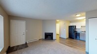 This single-floor space is designed with an open-concept layout that connects the living area to the kitchen and beyond. The living room is grounded by wall-to-wall carpeting and features a classic fireplace with a black surround, serving as a central focal point. Neutral beige walls and a textured popcorn ceiling contribute to a clean, understated aesthetic throughout. A baseboard heater runs along the wall, providing efficient supplemental warmth. A white entry door is positioned to one side of the living area, offering a clear and functional entryway. The flooring transitions from carpet in the living space to smooth hardwood-style planks as it extends into the kitchen and hallway areas. The kitchen is partially visible and includes white upper cabinetry and a black refrigerator, suggesting a functional and straightforward layout. A hallway leads further into the unit, hinting at additional rooms beyond the main living area. Recessed and flush-mount ceiling lighting provides adequate illumination throughout. Overall, the space presents a practical and well-organized interior with a neutral palette suited to a variety of furnishing styles.