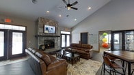 This single-floor space is thoughtfully designed to foster a warm and inviting atmosphere for gathering and relaxation. The centerpiece of the main lounge area is a striking floor-to-ceiling stone fireplace, which anchors the room with a sense of rustic elegance. A mounted flat-screen television above the fireplace adds a modern, functional touch. The seating arrangement features rich brown leather sofas and chairs positioned around a wooden coffee table atop a patterned area rug, creating a comfortable and well-appointed social hub. Vaulted ceilings rise overhead, enhancing the sense of openness, while recessed lighting and a ceiling fan with integrated lighting provide both ambiance and practicality. Natural light filters into the space through glass French doors at the rear, offering a view to the outdoors. A decorative wall feature displaying an inspirational "home" definition adds a personalized, community-focused touch. An adjacent room, partially visible through a doorway, hints at additional activity space within the property. The overall design balances casual comfort with polished finishes, making it well-suited for community events, social gatherings, or quiet relaxation.