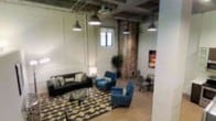 This single-floor space masterfully combines industrial character with contemporary comfort. The open-concept layout is anchored by an exposed ceiling with visible ductwork and pendant lighting, which casts a warm, inviting glow throughout. A striking exposed brick column serves as a natural focal point, adding raw texture and historic charm that contrasts beautifully with the clean white walls.

The lounge area is generously proportioned, featuring a dark leather sofa, teal accent chairs, and a bold geometric patterned area rug in earth tones that defines the seating zone. The furniture arrangement encourages both relaxed conversation and casual gatherings. Light wood flooring runs throughout, adding warmth and continuity to the space.

Natural light filters in through a window, complemented by the industrial-style pendant lights overhead. Decorative touches include a vibrant piece of wall art and a tall indoor plant, which introduce color and life to the interior. Additional seating is arranged toward the perimeter, offering flexible use of the space. Overall, the atmosphere is open, modern, and welcoming, with a distinctive character rooted in its industrial aesthetic and curated design elements.