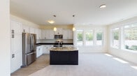 This single-floor residence is designed with an open, flowing layout that seamlessly connects the kitchen and living areas. The kitchen is a standout feature, showcasing white shaker-style cabinetry with dark countertops, a striking dark-base island topped with granite and fitted with a sink, and stainless steel appliances including a refrigerator, range, and built-in microwave. Pendant lighting hangs above the island, complementing the recessed ceiling fixtures throughout. Light wood-look flooring anchors the kitchen, transitioning to plush neutral carpet in the adjacent living and bedroom areas.

Large multi-pane windows are found throughout the home, inviting generous natural light and offering outdoor views. The bedrooms are comfortably sized with soft carpet flooring and ample window placement. A spacious walk-in closet provides extensive wire shelving and hanging storage. The bathrooms are well-appointed; one features a dark vanity with a white countertop and matte black hardware, while another includes a double-sink vanity with chrome fixtures and tile flooring. The overall palette of soft neutrals and contrasting dark accents creates a clean, modern aesthetic throughout.