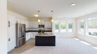 This single-floor residence is designed with an open, flowing layout that seamlessly connects the kitchen and living areas. The kitchen is a standout feature, showcasing white shaker-style cabinetry with dark countertops, a striking dark-base island topped with granite and fitted with a sink, and stainless steel appliances including a refrigerator, range, and built-in microwave. Pendant lighting hangs above the island, complementing the recessed ceiling fixtures throughout. Light wood-look flooring anchors the kitchen, transitioning to plush neutral carpet in the adjacent living and bedroom areas.

Large multi-pane windows are found throughout the home, inviting generous natural light and offering outdoor views. The bedrooms are comfortably sized with soft carpet flooring and ample window placement. A spacious walk-in closet provides extensive wire shelving and hanging storage. The bathrooms are well-appointed; one features a dark vanity with a white countertop and matte black hardware, while another includes a double-sink vanity with chrome fixtures and tile flooring. The overall palette of soft neutrals and contrasting dark accents creates a clean, modern aesthetic throughout.