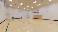 This single-level indoor basketball court offers a dedicated, full-featured athletic space designed for serious play and recreation. The court is finished with a classic light-toned hardwood floor, complete with regulation court markings in red — including the key, free-throw line, and center circle — providing an authentic playing experience. A wall-mounted basketball hoop with a clear glass backboard is positioned at one end, supported by a sturdy overhead arm system. The perimeter of the floor is bordered by a clean black edge, clearly delineating the playing surface. The walls are finished in crisp white, giving the space a clean, open feel, and branded court signage is displayed prominently on one wall. Folded bleacher-style seating is stored neatly along one side wall, offering the option to accommodate spectators. The ceiling is elevated to a generous height, well-suited for unobstructed gameplay, and is outfitted with a grid of recessed rectangular panel lights that deliver consistent, even illumination throughout the court. Ventilation is provided via ceiling-mounted vents. Overall, the space is clean, functional, and purpose-built for basketball, making it an excellent amenity for athletic and community use.