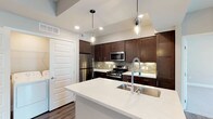 This single-floor space showcases a thoughtfully designed, contemporary kitchen that serves as its centerpiece. The kitchen is anchored by a generous center island topped with crisp white quartz countertops, offering ample workspace and a built-in stainless steel sink with a sleek, arch-neck faucet. Rich, dark espresso cabinetry lines the walls from floor to ceiling, providing substantial storage while lending a bold, sophisticated contrast to the light countertops. A decorative tile backsplash in neutral tones adds texture and visual interest between the upper and lower cabinets. The kitchen is fully equipped with stainless steel appliances, including a gas range, built-in microwave, and a French door refrigerator, catering to both everyday cooking and entertaining needs. Overhead, stylish cylindrical pendant lights hang above the island, complementing the recessed lighting that illuminates the space evenly. Warm-toned hardwood flooring runs throughout, grounding the modern aesthetic with a natural element. The layout is open and functional, with clean lines and a neutral color palette that creates a bright, welcoming atmosphere throughout the space.