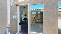 This single-story fitness center is designed to support a comprehensive workout experience in a bright, welcoming environment. The exterior features a stucco facade with large glass windows and a gated entry corridor, providing a secure and accessible entrance to the facility.

Inside, the gym boasts soaring ceilings that amplify the sense of openness throughout the space. A wall of tall, multi-pane windows along one side floods the interior with abundant natural light while offering pleasant views of surrounding palm trees and landscaping. Additional clerestory windows along upper walls further enhance the airy atmosphere.

The fitness floor is organized to accommodate a full range of training modalities. Cardio equipment includes multiple treadmills and stationary bikes positioned to take advantage of the scenic window views. The strength training area features a cable crossover machine, adjustable benches, and a full rack of dumbbells for free weight exercises. Several weight machines and specialty benches are arranged throughout the space for targeted muscle training.

Full-length mirrors line one wall, both expanding the visual depth of the room and aiding proper exercise form. A wall-mounted flat-screen television provides entertainment during workouts. The carpeted flooring offers comfort underfoot, while informational workout posters and safety signage are displayed throughout, making this a functional and well-appointed fitness facility.