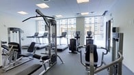 This fitness center is designed to support a comprehensive workout experience within a bright, modern environment. The space is outfitted with a variety of Life Fitness cardio equipment, including multiple treadmills and elliptical trainers, all positioned along a wall of large floor-to-ceiling windows that flood the room with natural light and offer views of the surrounding urban streetscape. The strength training area features a multi-station cable machine alongside additional resistance equipment, providing users with a range of exercise options. The room is finished with a drop ceiling featuring recessed panel lighting, which complements the natural light and ensures the space remains well-lit throughout the day. Dark rubberized flooring runs throughout, offering durability and safety underfoot. The layout is thoughtfully organized to allow users to move freely between stations without congestion. The overall ambiance is clean, functional, and contemporary, reflecting a professional fitness environment suited to a wide range of fitness routines.