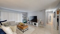 Located at Executive Center Drive in West Palm Beach, Florida, this single-floor residence offers a thoughtfully arranged layout encompassing a living room, dining area, kitchen, and a generously sized primary bedroom. The living room is finished with light-toned tile flooring and soft gray walls, creating a clean, neutral backdrop. Large windows dressed with vertical blinds invite natural light and lend an open, airy feel to the space. Wall-mounted shelving adds functional display space without compromising the room's openness. Adjacent to the living area, a dedicated dining space features tile flooring that continues from the main living area, accented by a contemporary sputnik-style chandelier and framed abstract artwork on the walls, adding a touch of artistic character. The kitchen is outfitted with warm wood cabinetry, a mosaic tile backsplash, white countertops, and stainless steel appliances, providing a practical and efficient cooking environment. The primary bedroom offers a comfortable retreat with textured ceiling detail, tile flooring, and ample room for a full furniture arrangement including a closet. Neutral gray tones throughout the residence create a cohesive and calm ambiance well-suited to the West Palm Beach lifestyle.