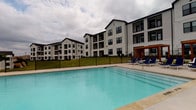 This outdoor pool area serves as a well-appointed recreational retreat within a residential community. The centerpiece is a generously sized swimming pool with clear, blue-tinted water, complemented by an adjacent spa for relaxation. The surrounding deck is finished in smooth concrete and offers ample space for lounging, furnished with multiple chaise lounge chairs and shaded by large patio umbrellas. A dedicated seating area features a comfortable sectional sofa and accent chairs, providing a social gathering space poolside. The pool is enclosed by a black iron security fence, ensuring safety while preserving open sightlines to the surrounding buildings and landscaped grounds. A poolside structure with warm wood-accented doors and stucco exterior provides access to the pool area and houses pool rules signage. Mature trees and ornamental plantings frame the perimeter, adding natural texture and shade. The multi-story residential buildings surrounding the pool feature a mix of brick and siding exteriors with private balconies overlooking the pool deck. The overall ambiance is open and resort-inspired, designed to encourage outdoor recreation and community interaction in a comfortable, well-maintained setting.