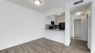 This single-floor space showcases a clean, modern design with an open-concept layout that connects the living area to the kitchen. The flooring throughout is a contemporary wood-look laminate in a warm grey-brown tone, providing a cohesive and stylish foundation across the space.

The kitchen is positioned behind a half-wall pass-through that doubles as a breakfast bar, creating a functional divide while maintaining visual openness to the main living area. White cabinetry lines the upper walls of the kitchen, offering ample storage, while a granite-patterned countertop adds a refined touch. A grey tile backsplash complements the counters and ties the aesthetic together. The kitchen is equipped with black appliances, including a refrigerator, microwave, and additional counter appliances, providing a bold contrast against the white cabinetry.

The walls throughout are painted a crisp white, maximizing the sense of brightness and space. Flush-mount ceiling lighting provides even illumination across both the kitchen and living areas. A hallway visible in the background suggests additional rooms beyond, rounding out the functional layout of this well-appointed space.