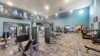 Located at 4600 Portofino Way in West Palm Beach, Florida, this two-story fitness center offers a spacious and well-appointed workout facility designed to accommodate a wide range of exercise needs. The ground floor welcomes visitors with soaring ceilings and soft blue accent walls, creating an open and energizing atmosphere. Patterned blue-and-tan carpeting runs throughout both levels, adding a contemporary aesthetic to the space. The main floor is generously outfitted with a broad selection of strength training machines, free weights, benches, and dedicated open floor areas, all arranged to maximize circulation and usability. Large windows and glass entry doors allow natural light to flood the interior while offering views to the outdoor surroundings. Multiple wall-mounted flat-screen televisions are positioned throughout, keeping members entertained during workouts. A mezzanine-level second floor, accessible via an interior staircase and framed by a metal railing system, houses additional cardio equipment including ellipticals and treadmills, providing an elevated vantage point over the main floor below. A boldly graphic striped mural decorates the upper level walls, contributing a dynamic visual element. Ceiling fans and recessed lighting ensure comfort throughout. The facility also features a mirrored wall section that amplifies the sense of space and supports technique-focused training.