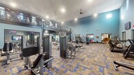 Located at 4600 Portofino Way in West Palm Beach, Florida, this two-story fitness center offers a spacious and well-appointed workout facility designed to accommodate a wide range of exercise needs. The ground floor welcomes visitors with soaring ceilings and soft blue accent walls, creating an open and energizing atmosphere. Patterned blue-and-tan carpeting runs throughout both levels, adding a contemporary aesthetic to the space. The main floor is generously outfitted with a broad selection of strength training machines, free weights, benches, and dedicated open floor areas, all arranged to maximize circulation and usability. Large windows and glass entry doors allow natural light to flood the interior while offering views to the outdoor surroundings. Multiple wall-mounted flat-screen televisions are positioned throughout, keeping members entertained during workouts. A mezzanine-level second floor, accessible via an interior staircase and framed by a metal railing system, houses additional cardio equipment including ellipticals and treadmills, providing an elevated vantage point over the main floor below. A boldly graphic striped mural decorates the upper level walls, contributing a dynamic visual element. Ceiling fans and recessed lighting ensure comfort throughout. The facility also features a mirrored wall section that amplifies the sense of space and supports technique-focused training.
