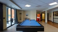 Located at 646 W Bay Street in Savannah, Georgia, this single-floor amenity space offers residents a comfortable and casual recreational area designed for leisure and social interaction. The room is anchored by a full-size billiards table with a vibrant blue felt surface, positioned prominently in the center of the open layout and ready for use with cue sticks mounted nearby. A drop-panel ceiling with recessed lighting provides consistent, even illumination throughout the space, while large windows along one wall allow natural light to filter in and offer views of the surrounding exterior. The flooring is finished in neutral tile, contributing to a clean and low-maintenance environment. A water fountain is conveniently accessible along the interior wall, and a Coca-Cola vending machine adds an easy refreshment option without leaving the floor. The open layout allows for easy movement throughout the space, with a hallway visible in the background suggesting connectivity to additional areas of the building. The overall ambiance is relaxed and functional, making it a practical gathering spot for social activities and everyday recreation.