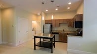 This single-floor residence offers a clean, modern aesthetic with a thoughtfully designed layout. The kitchen serves as the centerpiece of the space, showcasing warm honey-toned cabinetry that provides ample storage, complemented by granite countertops with subtle veining. Stainless steel appliances — including a full-size refrigerator, range, dishwasher, and built-in microwave — lend a sleek, contemporary feel. A freestanding kitchen island with a stainless steel top and lower storage shelf adds both functionality and a natural gathering point within the space. Overhead, a combination of recessed lighting and elegant pendant lights illuminate the kitchen, creating a warm and inviting ambiance. The open layout allows for easy flow between the kitchen and adjacent living areas. Light-toned flooring runs throughout, enhancing the sense of openness and brightness. A dedicated laundry area is conveniently tucked nearby, adding practical value to the home. The overall design balances modern finishes with everyday comfort, making this a highly functional and visually cohesive living space.