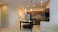 This single-floor residence offers a clean, modern aesthetic with a thoughtfully designed layout. The kitchen serves as the centerpiece of the space, showcasing warm honey-toned cabinetry that provides ample storage, complemented by granite countertops with subtle veining. Stainless steel appliances — including a full-size refrigerator, range, dishwasher, and built-in microwave — lend a sleek, contemporary feel. A freestanding kitchen island with a stainless steel top and lower storage shelf adds both functionality and a natural gathering point within the space. Overhead, a combination of recessed lighting and elegant pendant lights illuminate the kitchen, creating a warm and inviting ambiance. The open layout allows for easy flow between the kitchen and adjacent living areas. Light-toned flooring runs throughout, enhancing the sense of openness and brightness. A dedicated laundry area is conveniently tucked nearby, adding practical value to the home. The overall design balances modern finishes with everyday comfort, making this a highly functional and visually cohesive living space.