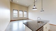 Located at 200 E First St in downtown Flint, MI, this two-floor loft-style unit offers a striking blend of industrial and modern design across a generous layout divided between two levels. The main floor impresses immediately with soaring ceilings that amplify the open, airy atmosphere, while polished concrete floors lend an industrial character throughout. A bank of large, black-framed windows floods the living area with natural light and frames views of the surrounding cityscape. The well-appointed kitchen is a standout feature, centered around a substantial island with a dark granite countertop and an undermount sink, illuminated by stylish pendant lights. White shaker cabinetry lines the walls alongside stainless steel appliances, including a refrigerator, range, and microwave, complemented by a tile backsplash and additional granite counter space. The bathroom is finished with clean subway tile walls, large-format floor tile, a wood-framed mirror, and a floating vanity with a marble-style countertop. An open staircase with a sleek cable railing leads to the upper loft level, where carpeted flooring, recessed lighting, and a built-in closet create a comfortable, versatile space. The loft overlooks the main floor below through an open railing, reinforcing the unit's dramatic vertical scale.