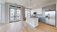 Located at 838 Morrison Dr in Charleston's vibrant 29403 district, this modern single-floor residence showcases a clean, contemporary design with light hardwood flooring and neutral tones carried throughout. The open-concept kitchen serves as the heart of the home, anchored by a large center island with a quartz countertop, an undermount sink with matte black fixtures, and elegant pendant lighting with gold-toned chains. Two-toned cabinetry — white upper cabinets paired with slate-gray lowers — complements a hexagonal tile backsplash, while stainless steel appliances complete the functional design. French doors adjacent to the kitchen open to a private balcony, flooding the living area with natural light and offering outdoor views. A well-proportioned hallway with recessed lighting connects the living spaces to the private rooms, each featuring ceiling fans, soft gray walls, and generous windows that bring in abundant daylight. One bedroom includes a spacious walk-in closet with built-in shelving. The bathroom is elegantly appointed with a soaking tub clad in large-format marble-style tile, a sleek floating vanity with ample drawer storage, and a wide mirror with contemporary lighting above the undermount sink.