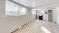 Located at 1136 Vine St in Liverpool, NY, unit D13 is a efficiently designed single-floor studio that makes smart use of its compact footprint. The main living area is bathed in natural light streaming through large windows fitted with horizontal blinds, which illuminate the light wood-tone laminate flooring that runs throughout the space. Clean white walls and a flush-mount ceiling fixture contribute to a bright, open feel. A baseboard heating unit and wall-mounted air conditioner ensure year-round comfort.

At one end of the living area, a kitchenette is neatly integrated into the layout, featuring a stainless steel refrigerator, dark cabinetry, and a sink — providing essential functionality within a minimal footprint. An electrical panel is conveniently positioned nearby.

Three white interior doors — including the main entry — line one wall, offering access to the bathroom and additional storage or closet space. The bathroom is finished in a clean, all-white palette with light wood-tone flooring consistent with the rest of the unit. It includes a vanity with cabinet storage, a toilet, and a full tub-shower combination with a white surround. The overall design of this unit is straightforward and functional, offering a well-maintained, move-in ready space.