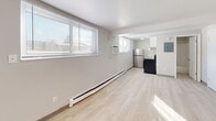 Located at 1136 Vine St in Liverpool, NY, unit D13 is a efficiently designed single-floor studio that makes smart use of its compact footprint. The main living area is bathed in natural light streaming through large windows fitted with horizontal blinds, which illuminate the light wood-tone laminate flooring that runs throughout the space. Clean white walls and a flush-mount ceiling fixture contribute to a bright, open feel. A baseboard heating unit and wall-mounted air conditioner ensure year-round comfort.

At one end of the living area, a kitchenette is neatly integrated into the layout, featuring a stainless steel refrigerator, dark cabinetry, and a sink — providing essential functionality within a minimal footprint. An electrical panel is conveniently positioned nearby.

Three white interior doors — including the main entry — line one wall, offering access to the bathroom and additional storage or closet space. The bathroom is finished in a clean, all-white palette with light wood-tone flooring consistent with the rest of the unit. It includes a vanity with cabinet storage, a toilet, and a full tub-shower combination with a white surround. The overall design of this unit is straightforward and functional, offering a well-maintained, move-in ready space.