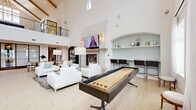 Located at 11023 Moonbug Ct in Charlotte, NC, this impressive living space is defined by its dramatic double-height ceilings and bright, airy atmosphere. The open-concept layout encompasses a generously sized living area, anchored by a striking stone fireplace with a mounted flat-screen TV and warm wall sconces on either side. Crisp white walls and light wood-look tile flooring flow seamlessly throughout, amplifying the sense of volume and openness. Large windows allow abundant natural light to pour in, enhancing the clean, modern aesthetic. A built-in arched display niche with seafoam-blue backing and open shelving adds an architectural focal point, accompanied by a bar-height counter with seating. The space is further enriched by a full-length shuffleboard table and a foosball table, making it highly suited for social gatherings. A centrally positioned wet bar island with quartz countertops, a tile backsplash, and barstool seating provides functional elegance. A grand staircase with dark metal railings and natural wood accents rises to an upper-level overlook balcony, adding vertical drama. A dining area with a long table rounds out this well-appointed, multi-functional gathering space.