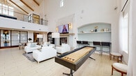 Located at 11023 Moonbug Ct in Charlotte, NC, this impressive living space is defined by its dramatic double-height ceilings and bright, airy atmosphere. The open-concept layout encompasses a generously sized living area, anchored by a striking stone fireplace with a mounted flat-screen TV and warm wall sconces on either side. Crisp white walls and light wood-look tile flooring flow seamlessly throughout, amplifying the sense of volume and openness. Large windows allow abundant natural light to pour in, enhancing the clean, modern aesthetic. A built-in arched display niche with seafoam-blue backing and open shelving adds an architectural focal point, accompanied by a bar-height counter with seating. The space is further enriched by a full-length shuffleboard table and a foosball table, making it highly suited for social gatherings. A centrally positioned wet bar island with quartz countertops, a tile backsplash, and barstool seating provides functional elegance. A grand staircase with dark metal railings and natural wood accents rises to an upper-level overlook balcony, adding vertical drama. A dining area with a long table rounds out this well-appointed, multi-functional gathering space.