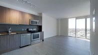 This single-floor space showcases a contemporary open-concept design that seamlessly integrates the kitchen and living areas into one cohesive environment. The kitchen is outfitted with stainless steel appliances, including an electric range, over-the-range microwave, and dishwasher, complemented by warm wood-tone cabinetry and a dark tile backsplash that adds visual depth. Track lighting on the ceiling provides directed illumination throughout the kitchen area. Light grey wood-style flooring extends throughout the entire space, contributing to a clean, modern aesthetic. The open living area is bright and airy, benefiting greatly from a dramatic floor-to-ceiling glass window that spans the far wall, flooding the interior with natural light and offering expansive elevated views of the surrounding cityscape and greenery. The white walls and neutral tones throughout create a minimalist atmosphere that feels fresh and versatile. The overall design emphasizes an uncluttered, functional layout with a strong connection between interior living and the outdoor environment beyond the glass.