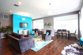 Located at 4133 Shoreline Cir in Virginia Beach, VA, this single-floor community gathering space combines modern design with welcoming warmth. Rich, dark hardwood flooring flows throughout, anchoring each zone with a cohesive and polished aesthetic. The main lounge area is a standout, featuring a bold teal accent wall that serves as a focal point alongside a wall-mounted flat-screen TV. Plush gray upholstered seating — including barrel chairs and a sofa — is arranged around a glass-top coffee table atop a vibrant ocean-inspired area rug, creating an inviting conversation space. Cowhide rugs add a touch of eclectic texture to the room. Natural light pours in through multiple large windows fitted with horizontal blinds, brightening the open layout. A dedicated dining area provides counter-height seating at a dark wood table, positioned beside a window with outdoor views. The kitchen is well-appointed with warm wood cabinetry, dark countertops, a refrigerator, microwave, and sink — well-suited for catering or communal use. Recessed lighting and pendant fixtures illuminate the space throughout, complemented by neutral gray walls that balance the vibrant teal accents. The overall ambiance is contemporary, energetic, and designed for comfortable social gatherings.