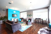 Located at 4133 Shoreline Cir in Virginia Beach, VA, this single-floor community gathering space combines modern design with welcoming warmth. Rich, dark hardwood flooring flows throughout, anchoring each zone with a cohesive and polished aesthetic. The main lounge area is a standout, featuring a bold teal accent wall that serves as a focal point alongside a wall-mounted flat-screen TV. Plush gray upholstered seating — including barrel chairs and a sofa — is arranged around a glass-top coffee table atop a vibrant ocean-inspired area rug, creating an inviting conversation space. Cowhide rugs add a touch of eclectic texture to the room. Natural light pours in through multiple large windows fitted with horizontal blinds, brightening the open layout. A dedicated dining area provides counter-height seating at a dark wood table, positioned beside a window with outdoor views. The kitchen is well-appointed with warm wood cabinetry, dark countertops, a refrigerator, microwave, and sink — well-suited for catering or communal use. Recessed lighting and pendant fixtures illuminate the space throughout, complemented by neutral gray walls that balance the vibrant teal accents. The overall ambiance is contemporary, energetic, and designed for comfortable social gatherings.