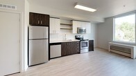 This contemporary single-floor unit offers a clean, modern aesthetic with a thoughtfully designed layout. The kitchen is a standout feature, showcasing a combination of white upper cabinetry and dark wood-grain lower cabinets with brushed silver hardware. It comes fully equipped with stainless steel appliances, including a refrigerator, electric range, over-the-range microwave, and dishwasher, all complemented by light quartz countertops and a single-basin sink with a modern faucet. Open shelving adds a functional design touch. Light wood-style vinyl flooring runs seamlessly throughout the unit, creating a bright and cohesive feel. The living area is generously proportioned, featuring clean white walls, ample wall outlets, and flush-mounted ceiling lighting that keeps the space feeling bright and airy. A secondary room provides flexible use as a bedroom or office, with a window allowing in natural light and a thermostat panel on the wall. The bathroom is finished with a granite-topped dark wood vanity, an undermount sink, tile-look vinyl flooring, and a full bathtub with chrome fixtures and grab bar. The overall palette of whites, grays, and warm wood tones gives the unit a polished, move-in-ready appearance.