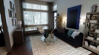 This single-floor space showcases a thoughtfully designed interior that blends modern aesthetics with a warm, inviting ambiance. The living area is a standout feature, highlighted by large floor-to-ceiling windows dressed with layered blinds and flowing curtains that allow abundant natural light to fill the room while offering privacy and light control. Dark hardwood flooring runs throughout, providing a rich contrast to the lighter elements in the space. The living area is anchored by a sleek black sofa adorned with an assortment of decorative pillows in teal, grey, and geometric patterns, complemented by a round glass-top coffee table with a sculptural metallic base. A bold blue canvas artwork commands attention on the feature wall, adding a vibrant pop of color to the neutral palette. An open metal-frame shelving unit serves both as a functional storage piece and a decorative accent. Soft ambient lighting from floor lamps adds warmth to the space during evening hours. A minimalist wireframe accent chair with a plush white faux-fur throw introduces an eclectic touch. The overall design sensibility is contemporary and curated, creating a space that feels both stylish and comfortable.