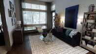 This single-floor space showcases a thoughtfully designed interior that blends modern aesthetics with a warm, inviting ambiance. The living area is a standout feature, highlighted by large floor-to-ceiling windows dressed with layered blinds and flowing curtains that allow abundant natural light to fill the room while offering privacy and light control. Dark hardwood flooring runs throughout, providing a rich contrast to the lighter elements in the space. The living area is anchored by a sleek black sofa adorned with an assortment of decorative pillows in teal, grey, and geometric patterns, complemented by a round glass-top coffee table with a sculptural metallic base. A bold blue canvas artwork commands attention on the feature wall, adding a vibrant pop of color to the neutral palette. An open metal-frame shelving unit serves both as a functional storage piece and a decorative accent. Soft ambient lighting from floor lamps adds warmth to the space during evening hours. A minimalist wireframe accent chair with a plush white faux-fur throw introduces an eclectic touch. The overall design sensibility is contemporary and curated, creating a space that feels both stylish and comfortable.