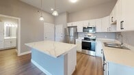 Located at The Elements in Rapid City, South Dakota, this well-appointed single-floor residence offers a thoughtfully designed layout with modern finishes throughout. The open-concept living area features durable wood-look flooring, neutral warm-toned walls, and a ceiling fan with lighting. Sliding glass doors lead to a private balcony, flooding the space with natural light and providing elevated outdoor views of the surrounding cityscape.

The kitchen is a standout feature, showcasing crisp white cabinetry, a large central island topped with a striking white quartz countertop with subtle veining, and a full suite of stainless steel appliances including a refrigerator, range, microwave, and dishwasher. Pendant lighting above the island and recessed ceiling lights enhance the bright, clean aesthetic.

The bedrooms are carpeted for comfort and benefit from generous natural light through large windows that frame views of the Rapid City surroundings. The bathroom is finished with a wide white vanity, a full tub-shower combination, wood-look flooring, and brushed nickel hardware and fixtures, delivering a clean and functional design. Exposed industrial-style ductwork in the living area adds a distinctive architectural touch, complementing the contemporary character of The Elements community.
