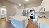 Located at The Elements in Rapid City, South Dakota, this well-appointed single-floor residence offers a thoughtfully designed layout with modern finishes throughout. The open-concept living area features durable wood-look flooring, neutral warm-toned walls, and a ceiling fan with lighting. Sliding glass doors lead to a private balcony, flooding the space with natural light and providing elevated outdoor views of the surrounding cityscape.

The kitchen is a standout feature, showcasing crisp white cabinetry, a large central island topped with a striking white quartz countertop with subtle veining, and a full suite of stainless steel appliances including a refrigerator, range, microwave, and dishwasher. Pendant lighting above the island and recessed ceiling lights enhance the bright, clean aesthetic.

The bedrooms are carpeted for comfort and benefit from generous natural light through large windows that frame views of the Rapid City surroundings. The bathroom is finished with a wide white vanity, a full tub-shower combination, wood-look flooring, and brushed nickel hardware and fixtures, delivering a clean and functional design. Exposed industrial-style ductwork in the living area adds a distinctive architectural touch, complementing the contemporary character of The Elements community.