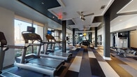This fitness center is designed to accommodate a comprehensive workout experience within a single, elongated space. The layout is thoughtfully organized, with distinct zones dedicated to cardio equipment and strength training. The cardio section lines one side of the room and features multiple treadmills, StairMaster climbers, stationary bikes, ellipticals, and a rowing machine, all positioned alongside floor-to-ceiling windows that flood the area with natural light and offer open views of the surrounding streetscape. The strength training zone toward the rear of the space is equipped with a full dumbbell rack, weight benches, cable machines, and functional training equipment. Large mirrors spanning the walls visually expand the space and allow users to monitor their form throughout their workout. The ceiling features a striking coffered design with recessed lighting and multiple ceiling fans, contributing to both the aesthetic and ventilation of the room. The flooring throughout consists of modular carpet tiles arranged in a bold geometric pattern of charcoal, taupe, and cream tones, adding visual interest while providing a durable, cushioned surface. A wall-mounted flat-screen television is positioned near the free weights area. A separate room with glass-panel doors provides additional training space.