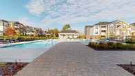 Located at 12509 Crown Park Ct in Charlotte, NC, the Steele Creek Rivergate Apartments clubhouse serves as the social hub of this well-appointed residential community. The single-story clubhouse is designed with a charming coastal-inspired aesthetic, combining shaker-style siding, brick detailing at the base, and large grid-pane windows that flood the interior with natural light. A welcoming entrance features a branded entry mat and elegant French doors framed by expansive white-trimmed windows, setting an inviting tone from the moment of arrival. Extending from the building is a generously sized covered porch supported by substantial columns, offering a comfortable outdoor living space furnished with wicker dining sets and decorative planters. This shaded veranda overlooks a sparkling resort-style swimming pool, surrounded by a broad, thoughtfully laid paver patio in a warm blend of earth tones. Lounge chairs line the pool deck, and manicured landscaping beds with seasonal plantings add a polished, natural touch throughout. The open-air setting provides sweeping views of the surrounding multi-story residential buildings, creating a strong sense of community. Outdoor televisions and additional seating areas enhance the entertainment potential of this versatile amenity space, making it ideal for both relaxing and socializing.