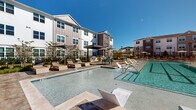 This outdoor amenity space serves as the centerpiece of a modern residential community, offering a resort-inspired retreat designed for relaxation and socializing. The expansive swimming pool is the focal point, featuring a shallow sun shelf lined with contemporary lounge chairs that allow residents to enjoy the water while basking in natural sunlight. The pool deck is finished with stylish paver stone surroundings, providing ample space for lounging with additional chaise chairs and market umbrellas positioned throughout.

A covered pergola structure anchors the entertainment area, housing an outdoor kitchen equipped with grills, making it well-suited for gatherings and cookouts. Adjacent to the pool, a second shallow water feature adds a playful, resort-like element to the space. Additional covered pergola seating areas with picnic-style tables extend the outdoor living space, draped with overhead string lights that create an inviting evening ambiance.

The community's multi-story residential buildings, constructed with a mix of white board-and-batten siding and warm brick accents, frame the pool area and lend a contemporary yet welcoming architectural character. Newly planted landscaping, ornamental lamp posts, and a wrought iron perimeter fence complete the polished, well-appointed outdoor environment.