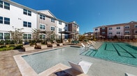 This outdoor amenity space serves as the centerpiece of a modern residential community, offering a resort-inspired retreat designed for relaxation and socializing. The expansive swimming pool is the focal point, featuring a shallow sun shelf lined with contemporary lounge chairs that allow residents to enjoy the water while basking in natural sunlight. The pool deck is finished with stylish paver stone surroundings, providing ample space for lounging with additional chaise chairs and market umbrellas positioned throughout.

A covered pergola structure anchors the entertainment area, housing an outdoor kitchen equipped with grills, making it well-suited for gatherings and cookouts. Adjacent to the pool, a second shallow water feature adds a playful, resort-like element to the space. Additional covered pergola seating areas with picnic-style tables extend the outdoor living space, draped with overhead string lights that create an inviting evening ambiance.

The community's multi-story residential buildings, constructed with a mix of white board-and-batten siding and warm brick accents, frame the pool area and lend a contemporary yet welcoming architectural character. Newly planted landscaping, ornamental lamp posts, and a wrought iron perimeter fence complete the polished, well-appointed outdoor environment.