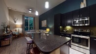 This thoughtfully designed single-floor residence combines contemporary style with functional living. The open-concept kitchen and living area serves as the heart of the home, anchored by a striking dark granite island with bar seating and pendant lighting. The kitchen is outfitted with dark cabinetry, a green tile backsplash, stainless steel appliances including a refrigerator, range, and built-in microwave, all set against rich hardwood-style flooring. A bold teal accent wall ties the kitchen seamlessly into the living room, where tall ceilings, a large window framing lush greenery, and warm wood floors create an inviting atmosphere. The bedrooms are serene and well-appointed, featuring plush carpeting, generous windows with outdoor views, and soft neutral tones. A versatile den or office space offers large windows overlooking tree canopy views, providing an ideal work-from-home environment. Two bathrooms feature dark vanity cabinetry with light countertops and large mirrors, with one including a full soaking tub and shower combination with classic tile surround. Throughout the residence, the interplay of dark and light tones, natural light, and greenery views lends the space a refined yet comfortable ambiance.