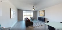 This single-floor, one-bedroom, one-bathroom residence is designed with a clean, contemporary aesthetic that prioritizes natural light and functional living. The living area serves as the centerpiece of the space, featuring floor-to-ceiling sliding glass doors that flood the room with daylight and offer open views of the surrounding landscape. Light-toned hardwood-style flooring runs throughout, complementing the neutral gray wall palette and contributing to a cohesive, modern atmosphere.

A ceiling fan with a brushed chrome finish is centrally positioned overhead, providing both practical airflow and a subtle design accent. The living area comfortably accommodates a full seating arrangement, with ample room for a sofa, accent chair, glass coffee table, and a substantial storage console. A textured area rug anchors the seating zone and adds warmth to the space.

The layout is straightforward and efficient, making excellent use of the available square footage. White baseboards and clean wall transitions provide a polished finish throughout. Overall, the residence offers a bright, modern environment well-suited for comfortable everyday living.