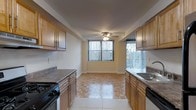 This single-floor residence offers a thoughtfully designed layout that balances functionality with a warm, inviting ambiance. The kitchen is a standout feature, showcasing rich honey-toned wood cabinetry that lines both sides of the galley-style space, providing generous storage and a classic aesthetic. Laminate countertops offer ample workspace, and the kitchen is equipped with a gas range and a built-in sink, making it well-suited for everyday cooking. The flooring transitions from tile in the kitchen area to a beautifully patterned parquet in the adjoining dining or living space, adding texture and visual interest underfoot. Large windows dressed with vertical blinds allow abundant natural light to fill the space, creating a bright and airy atmosphere throughout. A ceiling fan overhead in the adjoining area provides both comfort and additional illumination. The neutral wall tones complement the warm wood elements, lending the space a cohesive and welcoming character. Overall, this residence combines practical features with classic design details, making it a comfortable and functional living environment.