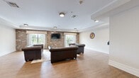 Located at 120 Chubb Ave, this single-floor living space offers a thoughtfully designed communal area that blends classic character with modern functionality. The living room is anchored by a striking exposed brick accent wall, which serves as a natural backdrop for a wall-mounted flat-screen television flanked by two large windows dressed with patterned valances. These windows invite generous natural light and offer pleasant outdoor views, enhancing the open and airy atmosphere of the room.

The flooring throughout is warm, light-toned hardwood, complemented by crisp white walls and elegant tray ceilings with crown molding that add architectural depth. Tufted Chesterfield-style leather sofas in rich brown provide a classic, inviting seating arrangement centered around the main television wall.

A secondary area features an additional Chesterfield loveseat, wall-mounted shelving for books or media, and vibrant wall art that adds a pop of color to the neutral palette. A small dining nook with a white table and mixed chairs offers a casual gathering spot.

A distinctive design highlight is the dark mosaic tile accent wall housing dual drinking fountains — a unique, utilitarian feature that adds texture and visual interest. Together, these elements create a warm, versatile, and welcoming space ideal for relaxation and social interaction.