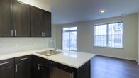 This single-floor residence showcases an open-concept layout that seamlessly connects the kitchen and living area, creating a cohesive and functional living environment. The kitchen is outfitted with dark espresso cabinetry equipped with sleek bar-pull hardware, contrasting elegantly against crisp white countertops. A peninsula with an undermount stainless steel sink and integrated dishwasher extends the workspace while visually anchoring the kitchen within the larger open floor plan. A subway tile backsplash adds a clean, classic finish to the cooking space.

The living area benefits from generous natural light streaming through multiple windows, including a large sliding glass door that opens to a private balcony with a metal railing — offering both an indoor-outdoor connection and added ventilation. An additional side window further brightens the space throughout the day. The flooring throughout features hardwood-style planks that add warmth and continuity across both the kitchen and living zones. Neutral, light gray walls complement the overall palette, lending the space a modern yet inviting ambiance. Recessed lighting provides consistent illumination throughout the interior.