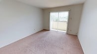 This single-floor residential space is designed with simplicity and functionality in mind, offering a clean, neutral palette that provides a versatile canvas for personalization. The living area is a standout feature, characterized by soft, plush carpeting in warm neutral tones that adds comfort underfoot and contributes to the room's welcoming ambiance. Smooth, light-colored walls complement the overall airy feel of the space.

A prominent sliding glass door serves as both a practical and aesthetic focal point, flooding the room with abundant natural light and offering a direct connection to an outdoor balcony. Vertical blinds provide flexible light control and privacy as needed. The balcony beyond hints at outdoor views of surrounding greenery and neighboring structures, offering a pleasant outdoor retreat.

The layout is open and unobstructed, allowing for flexible furniture arrangement to suit a variety of living styles. The clean lines of the architecture keep the focus on the sense of openness and light throughout. Overall, this space presents a bright, comfortable, and straightforward living environment, well-suited for those seeking a functional and easily adaptable home.