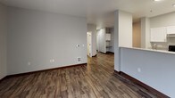 This single-floor residential unit offers a clean, modern interior with a cohesive neutral color palette and wood-look luxury vinyl plank flooring throughout the main living areas. The open living space is airy and functional, anchored by a ceiling fan with light fixture and seamless sightlines toward the kitchen. A half-wall partition provides a natural division between the living area and the galley-style kitchen, which features white shaker cabinetry, light stone countertops, a stainless steel sink, and a full suite of appliances including an electric range, refrigerator, and dishwasher. The bedrooms are carpeted and filled with natural light from large windows with horizontal blinds, and each is equipped with baseboard heating. The unit includes two bathrooms: a full bath with a soaking tub and shower combination, a white vanity with stone countertop, and wood-look flooring; and a second bathroom with a standalone walk-in shower, toilet, and matching flooring. Both bathrooms feature bright, clean finishes with brushed nickel hardware. The overall design emphasizes practicality and comfort, with consistent trim work, neutral gray walls, and a bright, move-in-ready aesthetic throughout.