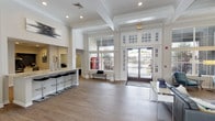 This single-floor community clubhouse offers a well-appointed gathering space designed for comfort and socializing. The entrance welcomes visitors with soaring coffered ceilings, crisp white millwork, and large floor-to-ceiling windows that flood the space with natural light and offer views of the surrounding outdoor areas. Wide-plank wood-style flooring flows throughout, tying together the various zones seamlessly.

The main lounge area serves as the heart of the space, anchored by a dramatic floor-to-ceiling tiled fireplace framed by white columns. Stylish seating, including teal velvet accent chairs and contemporary sofas, creates comfortable conversation areas. A bar-height counter with black barstools and granite-style surfaces provides additional gathering space.

The fully equipped kitchen features stainless steel appliances, a sleek electric range, a stainless range hood, granite countertops, white cabinetry, and a striking dark mosaic tile backsplash — ideal for hosting events. A beverage station adds convenience for residents.

A dedicated game room offers a foosball table, a leather sofa, and relaxed lounge seating beneath a skylight, making it an inviting retreat for recreation. Throughout the clubhouse, modern artwork and carefully chosen décor contribute to a polished yet approachable ambiance.