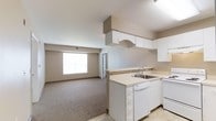 This single-floor residential unit offers a practical and comfortable layout designed for everyday living. The open-concept design seamlessly connects the kitchen and living area, creating a spacious and functional flow throughout the main space. The kitchen is well-appointed with white cabinetry, a light-toned countertop, a stainless steel sink, a dishwasher, and overhead cabinets that maximize storage. A decorative arched cabinet detail adds a subtle design touch above the counter. The kitchen transitions smoothly into the living area, which features wall-to-wall carpet in a neutral beige tone, contributing to a warm and quiet atmosphere. A large window allows generous natural light to fill the living space, brightening the room and enhancing its open feel. The walls throughout are finished in a soft, neutral beige palette that complements the overall understated aesthetic. The unit also includes a doorway leading to additional areas, suggesting a layout with further rooms beyond the main living space. Light tile flooring in the kitchen contrasts gently with the carpeted living area, clearly defining each functional zone. Overall, the space presents a clean, move-in ready environment with a straightforward and efficient design.
