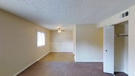 This single-floor residence at Iroquois Gardens offers a practical and comfortable layout with a welcoming interior. The main living area is generously proportioned, featuring warm-toned carpet flooring that transitions to hard flooring in an adjacent dining or secondary space. Soft yellow walls contribute to a bright, inviting atmosphere, complemented by natural light filtering in through a window fitted with blinds. A ceiling fan with a light fixture is centrally positioned, providing both air circulation and ambient lighting. A textured popcorn ceiling adds a classic character to the space. One wall in the adjoining area features a subtle striped wallpaper detail along the upper half, adding a touch of visual interest above the white chair rail molding. White baseboards and clean trim work run throughout, offering a crisp contrast to the warm wall tones. An open pass-through or partial wall delineates the living and adjacent areas while maintaining an open, connected feel. The overall ambiance is clean, neutral, and move-in ready, offering a straightforward and functional living environment.