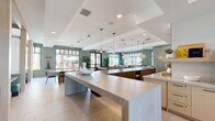 Located at 1464 Laurel Sound Ln in Sanford, Florida, this expansive single-floor community clubhouse offers a generously sized, thoughtfully designed space for socializing, working, and recreation. The interior is defined by soaring double-height ceilings in the entrance area, exposed wood beam accents, and large black-framed windows that flood the space with natural light and provide views of the surrounding community and outdoor greenery.

The open-plan layout flows seamlessly between distinct zones. A co-working area near the entrance features structural wood columns and ample desk space. Adjacent to it, a fully equipped coffee and refreshment bar offers light cabinetry, quartz countertops, and a commercial beverage station beneath a dramatic skylight. A long island with bar seating and pendant lighting anchors the central social hub, complemented by a large-screen TV and display shelving.

The dining and lounge areas are elegantly appointed, with leather banquette seating, statement pendant chandeliers, and floor-to-ceiling windows. A cozy nook with green upholstered benches and a dark wood table offers a more intimate retreat. The game room features a billiards table, potted fiddle-leaf fig trees, and direct access to the outdoor pool area through glass-paned French doors. Throughout, sage green accent walls, light stone tile flooring, and curated decor create a cohesive, contemporary Florida ambiance.