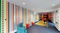 This dynamic entertainment space combines bold design with recreational amenities, creating an energetic and visually striking environment. The most prominent feature is the private bowling lane, finished with classic hardwood-style flooring and complete with a ball return rack stocked with colorful bowling balls. A retro neon "Lucky" sign glows above the lane, lending the space a playful, arcade-inspired character, while an overhead monitor enhances the bowling experience.

The adjacent lounge area provides comfortable seating, including a curved teal velvet chair complemented by a collection of colorful round ottomans in shades of orange, red, and lavender, arranged informally on plush grey carpeting. A large glass panel wall divides the lounge from a secondary room, maintaining visual openness throughout the space.

The most visually commanding element is the floor-to-ceiling multicolored vertical stripe wall mural, which spans the length of the room and introduces a lively, artistic focal point. Recessed ceiling lighting provides clean, even illumination throughout. Overall, the space balances retro-inspired aesthetics with modern design sensibilities, making it a highly functional and entertaining environment.